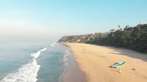 Golden-tropical-sandy-beach-with-moored-fishing-boats-and-tourists-out-walking-in-Varkala,-Kerala,-India---Aerial-Low-angle-Fly-over