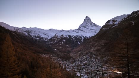 Day-to-night-timelapse-of-the-Matterhorn-as-seen-from-Zermatt,-Switzerland-as-the-stars-light-up-the-sky-along-with-the-city-lights