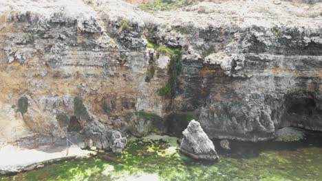 Small-waterfall-in-rocky-cliff-cascading-down-through-the-boulders-to-the-mediterranean-sea-in-Malta---Aerial-Fly-backwards-reveal-shot