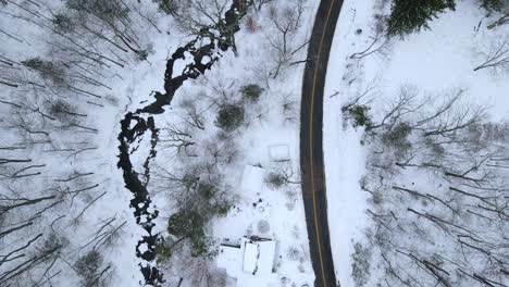 Birds-eye-view-of-a-snowy,-remote-mountain-road-with-a-stream-and-forest