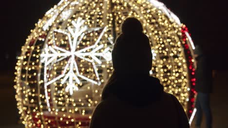 Woman-Standing-Against-Large-and-Bright-Christmas-Bauble-Decoration-at-Night