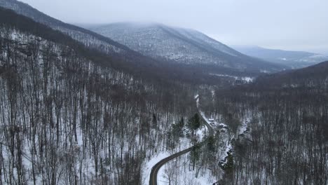Descending-on-a-snowy,-forested-mountain-valley-during-winter-with-mountains-and-a-remote-road-and-cloud-skies