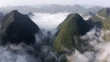 Montañas-Kársticas-Asiáticas-Que-Se-Elevan-Por-Encima-De-Las-Nubes-Y-Un-Pequeño-Pueblo-Chino,-Antena