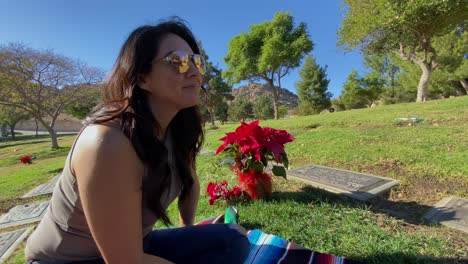 Close-up-of-hispanic-female-sitting-on-grass-at-cemetery-paying-her-respects,-smiling-on-a-clear-sunny-day
