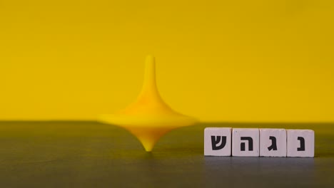 A-plastic-top-spins-and-wobbles-to-a-stop-beside-the-four-hebrew-letters-nun,-gimel,-heh-and-shin-shown-against-a-two-tone-background