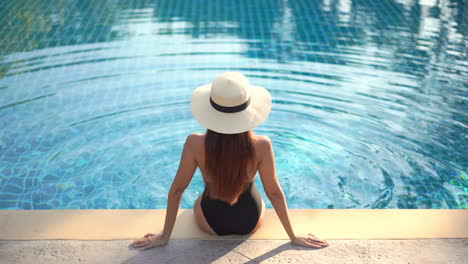 A-healthy,-fit-sexy-woman-sits-on-the-edge-of-a-swimming-pool-back-to-the-camera-as-she-admires-the-view