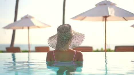 A-woman-wearing-a-straw-sun-hat,-while-relaxing-in-a-resort,-swimming-pool-looks-out-the-ocean-beyond