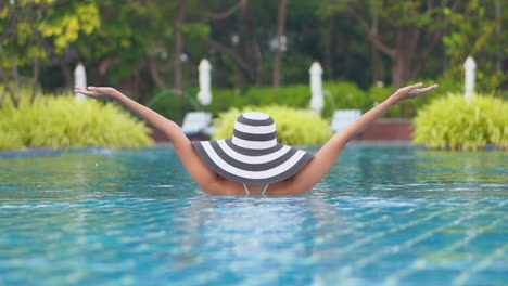 Back-view-of-female-tourist-standing-half-submerged-in-the-swimming-pool-wearing-a-big-black-and-white-hat,-rising-her-arms-above-her-head,-enjoying-her-tropical-holidays