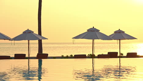 Eine-Schöne-Aussicht-Auf-Einen-Romantischen-Sonnenuntergang-An-Einem-Klaren-Goldenen-Himmel-Und-Einem-Ruhigen-Ozean-Im-Hintergrund-Mit-Leeren-Stühlen,-Sonnenschirmen-Und-Einem-Swimmingpool-Im-Vordergrund