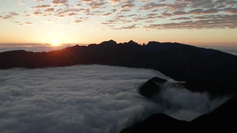 Sea-of-clouds-flows-between-dark-mountains-during-colorful-sunrise,-aerial