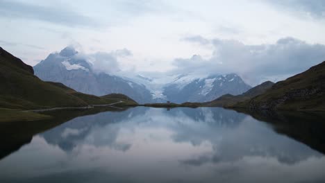 Day-to-night-timelapse-of-lake-Bachalpsee-in-Grindelwald,-Switzerland-with-a-view-of-alpine-peaks,-glaciers-and-stars-reflected-on-the-lake's-surface