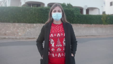 Young-adult-wearing-a-Christmas-sweater-putting-on-a-surgical-mask-in-the-street-and-looking-at-the-camera