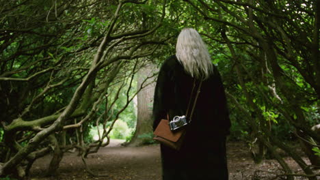 Young-blond-photographer-hipster-girl-woman-walking-through-a-magical-story-forest-with-scary-trees-with-vintage-bag-and-camera-Fujifilm-old-photographic-camera-capturing-adventure-European-summer