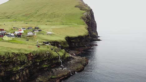 Aerial-of-small-village-along-the-cliffside-of-small-mountain-with-waterfalls-and-rivers-in-the-lush-landscape-covered-in-grass-next-to-the-calm-North-Atlantic-ocean