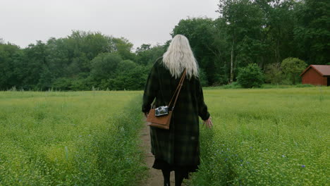 Blonde-cute-German-girl-walking-across-a-field-of-flowers-in-a-black-coat-vintage-bag-and-camera-towards-a-forest-on-misty-cloudy-Scandinavian-spring-touching-with-hands-wanderlust-exploring-adventure