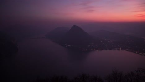 Sunset-timelapse-of-Lugano-city-and-lake-from-Monte-Bre,-Switzerland-with-an-orange-sky-turning-blue-as-the-night-sky-lights-up-by-the-city-lights