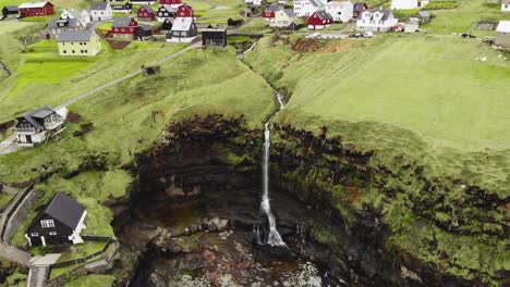 Aerial-of-small-village-on-the-cliffside-with-lush-grass-and-small-rivers-and-waterfalls-falling-into-the-calm-North-Atlantic-ocean-in-the-fjord-at-the-Faroe-Islands