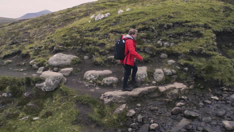 Young-man-with-red-jacket-and-a-black-backpack-is-crossing-a-small-creek-river-on-the-mountain-side-on-his-hike-in-slow-motion-on-the-Faroe-Islands