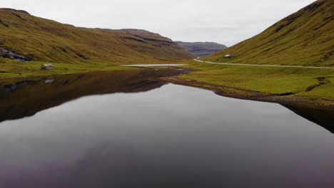 Aerial-low-angle-with-high-speed-over-beautiful-lake-with-calm-reflective-water-in-the-mountains-next-to-a-road-on-the-Faroe-Islands