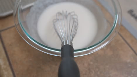 Whisk-in-bowl-of-white-icing-glaze-on-countertop,-Overhead-Detail-Approach