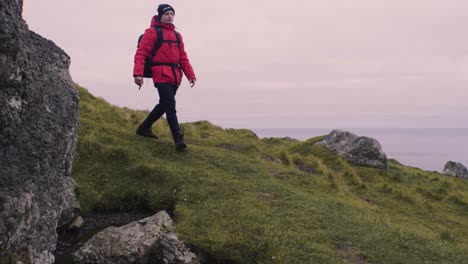 Man-in-red-jacket-and-with-a-backpack-on-hikes-through-the-green-hills-and-big-rocks-on-the-beautiful-mountain-trek-on-The-Faroe-Islands