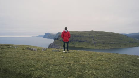 Man-in-red-jacket-standing-om-a-hill-and-staring-out-on-the-amazing-landscape-view-with-fjords,-mountains-and-cliffs-on-a-cloudy-day-on-Vagar-on-The-Faroe-Islands