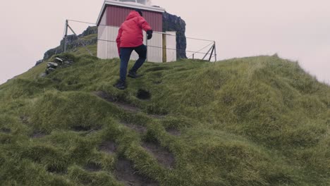 Man-in-red-jacket-hikes-up-a-green-mountain-and-gets-to-the-lighthouse,-Kalsoy-at-the-top-on-the-Faroe-Islands-on-a-cloudy-day
