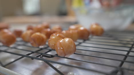 Woman-places-fresh-donut-holes-dripping-in-glaze-on-cooling-rack,-Close-Up