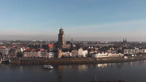 Ferryboat-crossing-the-river-IJssel-with-backdrop-of-Dutch-Hanseatic-medieval-city-of-Deventer-in-The-Netherlands-reflecting-in-the-water-revealing-the-quay-and-scaffolding-with-a-Christmas-tree