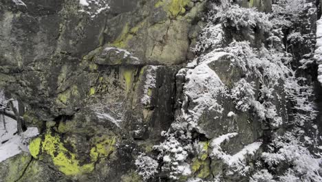 Rising-up-a-moss-covered-stone-formation-as-snow-gently-falls,-Rocher-de-Bilisse,-aerial-closeup