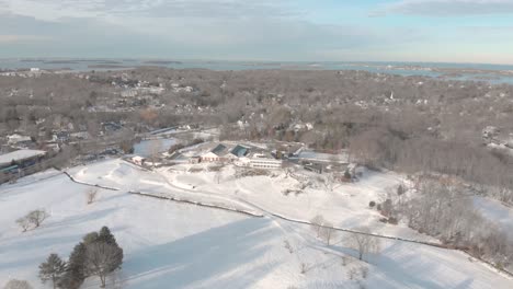 At-an-elevated-height-and-view-straight-ahead,-the-drone-view-of-a-snow-covered-golf-course-with-the-clubhouse-close-and-Boston-in-the-distance-is-mesmerizing