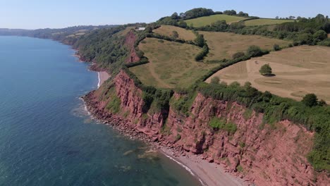 Epic-drone-shot-of-red-clay-coastline,-rocky-shore,-clear-blue-ocean-water-and-green-rural-landscape-on-top