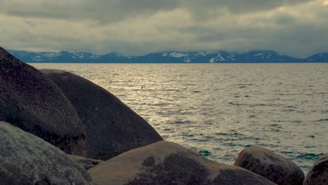 Large-boulder-rock-formations-on-the-side-of-South-Lake-Tahoe-with-snow-capped-mountains-in-the-background-surrounded-by-clouds---Sand-Harbor