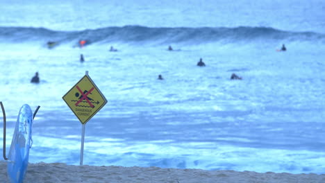 Dangerous-Current-Warning-Sign-In-Bronte-Beach-With-Surfers-In-Background-In-NSW,-Australia