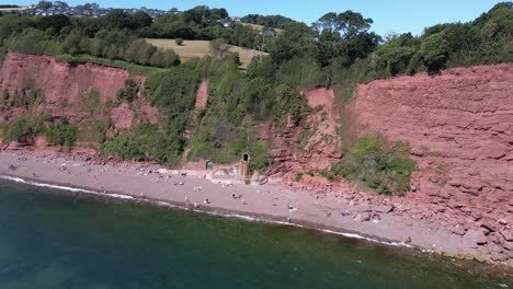 Ascending-aerial-backwards-shot-of-people-resting-on-ness-cove-beach-with-red-cliff-in-background-and-rural-city-landscape-on-top
