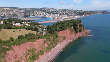 Aerial-flight-over-ness-cove-beach,-buildings-on-top-and-beautiful-Teignmouth-Port-in-background