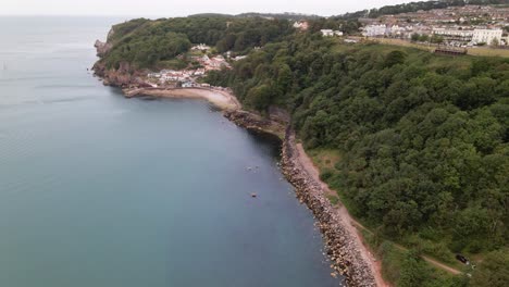 Aerial-circling-shot-of-stony-famous-Babbacombe-Beach-with-green-cliffs-and-Torquay-Town-on-top-of-mountain