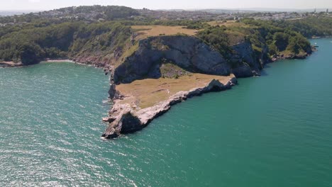 Aerial-pan-shot-of-famous-Long-Quarry-Point-on-high-hill-surrounded-by-green-blue-ocean-water-in-sun