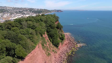 Aerial-flight-along-red-cliff-with-trees-on-top-and-beautiful-Shaldon-city-in-background
