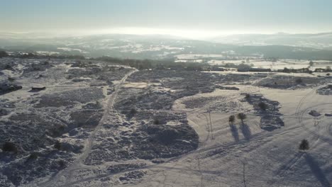 Snow-covered-rural-winter-countryside-track-footprint-shadows-terrain-aerial-view-rising-tilt-down