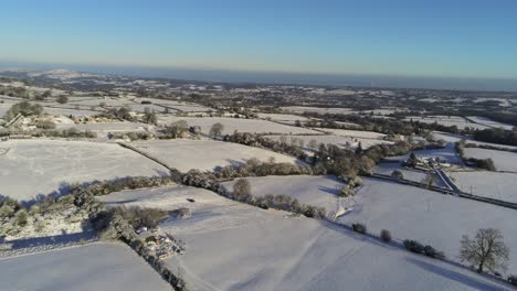Cold-snowy-winter-British-patchwork-farmland-countryside-rural-scene-aerial-rising-at-sunrise