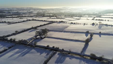 Cold-snowy-winter-British-patchwork-farmland-countryside-rural-scene-aerial-at-sunrise-cinematic-orbit-left