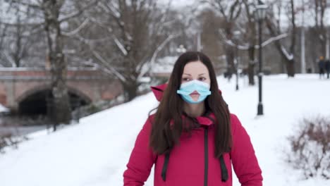 Woman-Standing-In-Snowy-City-Park-Wearing-A-Face-Mask-With-Hole-During-Winter