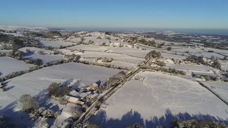 Cold-snowy-winter-British-patchwork-farmland-countryside-rural-scene-aerial-at-sunrise-tilting-down