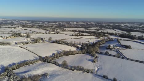 Cold-snowy-winter-British-patchwork-farmland-countryside-rural-scene-aerial-at-sunrise-cinematic-pan-right
