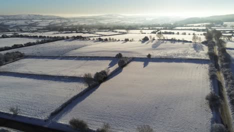 Cold-snowy-winter-British-patchwork-farmland-countryside-rural-scene-aerial-at-sunrise-flyover-slow
