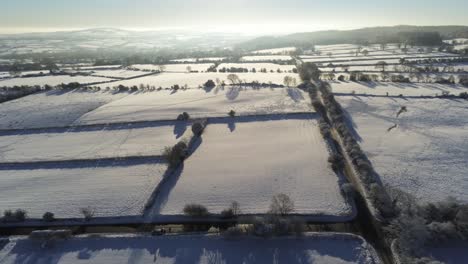 Cold-snowy-winter-British-patchwork-farmland-countryside-rural-scene-aerial-at-sunrise-forward-descent-right