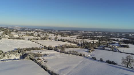 Cold-snowy-winter-British-patchwork-farmland-countryside-rural-scene-aerial-slow-rising-tilt-down-at-sunrise