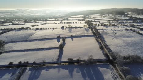 Cold-snowy-winter-British-patchwork-farmland-countryside-rural-scene-aerial-at-sunrise-slow-descend-forwards