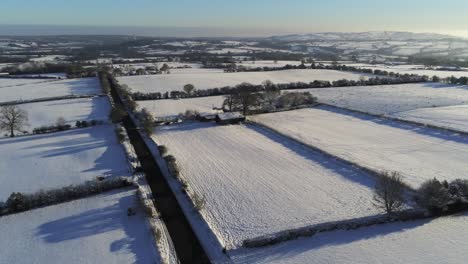 Cold-snowy-winter-British-patchwork-farmland-countryside-rural-scene-aerial-at-sunrise-left-fast-pan